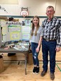 Callia - a girl with long brown hair in blue jeans and gray t-shirt standing next to her grandpa - a man with glasses and long mustache - at an AgrAbility booth
