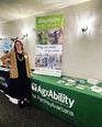 Kendra Martin in dark dress and tan sweater standing next to AgrAbility for Pennsylvanians table with AgrAbility literature on it and an AgrAbility banner on the left end of the table