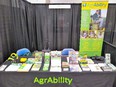 Table with AgrAbility tablecloth on it & brochures & assistive technology displayed. AgrAbility banner in background with black curtains around booth