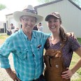 Person on left with Stetson hat - glasses - blue checked shirt with arm around woman in brown coveralls & baseball cap with pole barn-shed in background