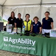 5 women standing behind green-draped AgrAbility for Pennsylvanians display table in a white tent.