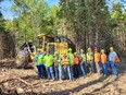 A group of logging students in florescent clothes & hard hats standing in front of trees on a section of denuded land with a large yellow logging machine behind them