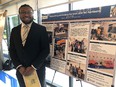 Young African American man in sport jacket & tie wearing glasses & standing next to a poster titled NETWORKING & HELPING FARMERS WITH NC AGRABILITY