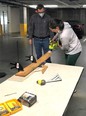 A man and a woman using hand power tools to cut a board in what looks like a basement parking garage