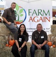 A woman sitting in between 2 men on hay bales with small pumpkins beside them & a Farm Again banner behind them