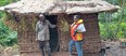 African mud hut with plastic-covered grass roof & a man standing in doorway speaking with woman who is wearing orange & yellow life vest