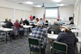 Classroom with people seated at rectangular white tables listening to speaker at front with PowerPoint on screen