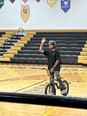Man wearing helmet straddling bicycle and waving to crowd in gymnasium