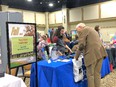 Large room with many booths in it & Abbie Spackman at AgrAbility for Pennsylvanians behind blue-draped table talking with white-haired man in front of her