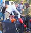 Black-draped table in foreground with powered yard equipment on it and man showing the tools to people around the table.