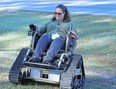 Woman in green top and blue pants driving a Trackchair in the grass next to a paved road.