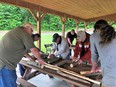 Men around a table in a picnic shelter working with small logs about 2 inches in diameter by 16 inches long