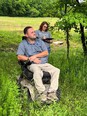 Man in a wheelchair outdoors in a field with a woman behind him writing on a clipboard
