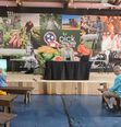 TN Region  2N Coordinator speaking on platform with large murals of farmers at work in background At TN State Fair