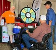 NE AgrAbility booth with spinning-wheel learning device on table and a man in a motorized wheelchair in the foreground