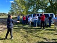 Tour group standing in a field in front of a white van with trees in the background & a woman speaking to them