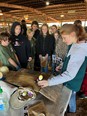 A group of youth in a large farm building watching a demonstration on a table.