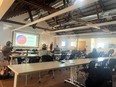 People at long tables in a large room listening to a speaker with a PowerPoint presentation