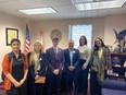 2 men in center and 4 women standing in office with 2 flags behind them