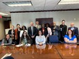 14 people standing or sitting behind a large wood table and smiling at the camera