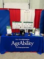 Picture of AgrAbility for Pennsylvanians booth with small AT items & brochures on blue-draped table with redd & white curtains as backdrop