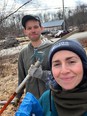 A man and woman with a pruning saw outside with a gray barn and woods in the background