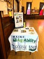 Maine AgrAbility display table with literature and a small pop-up banner on the table in a large room.