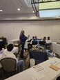 Woman in a large room presenting items from a display table to a group of people