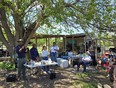 A group of people under a large tree listening to a man speak with what looks like a concession stand behind them