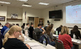 Classroom full of people seated at long desks listening to speaker with PowerPoint presentation