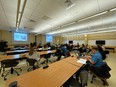 Joe Ricker at the front of a large classroom with long tables and a group of people sitting at them
