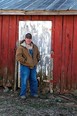 Farmer - Jason - in overalls and brown coat standing in front of red shed with a white door
