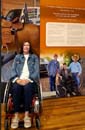Woman in a wheelchair in front of an AgrAbility exhibit at the NM Farm & Ranch Heritage Museum