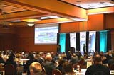 A group of people seated at round tables in a large conference room listening to 2 presenters on stage