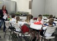A young woman speaking to a group of at least 8 children seated at long white tables in a room that has many potted plants.