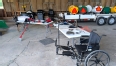 2 long white tables with AT items on display and a wheelchair at one end.