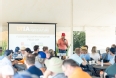 A man - Dr. Hatcher - using PowerPoint while speaking to a group of people seated at long tables under a large tent