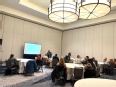 People seated at tables in a large conference room listening to a speaker and watching a PowerPoint.